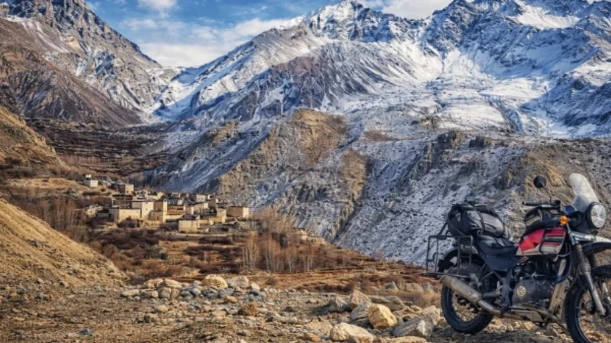 Adventure motorcycle parked on a rugged Himalayan mountain road with snow-capped peaks and a remote village in Nepal