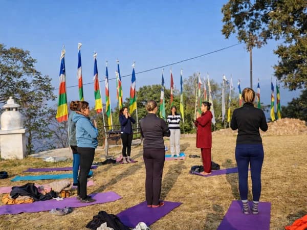 People practicing yoga outdoors on a hillside with colorful prayer flags in the background.